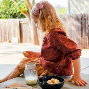 May include: A young girl with blonde hair wearing a brown shirt and a pink bow sits on a wooden surface with a glass of milk and a bowl of cookies.