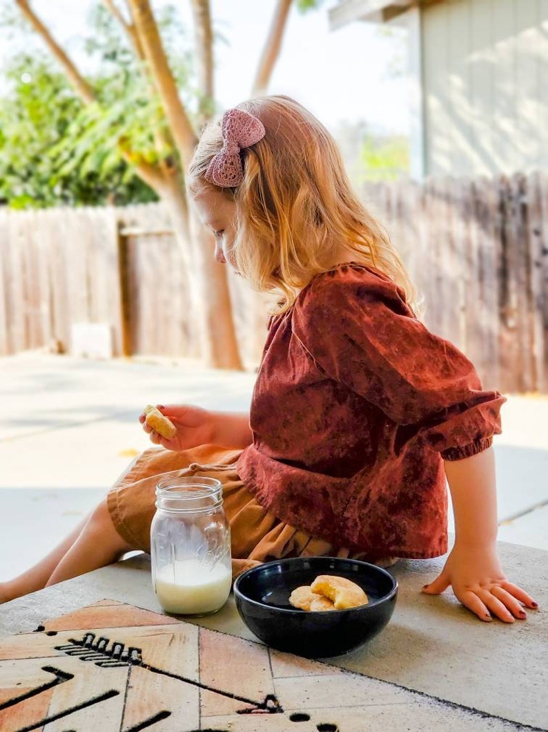 May include: A young girl with blonde hair wearing a brown shirt and a pink bow sits on a wooden surface with a glass of milk and a bowl of cookies.