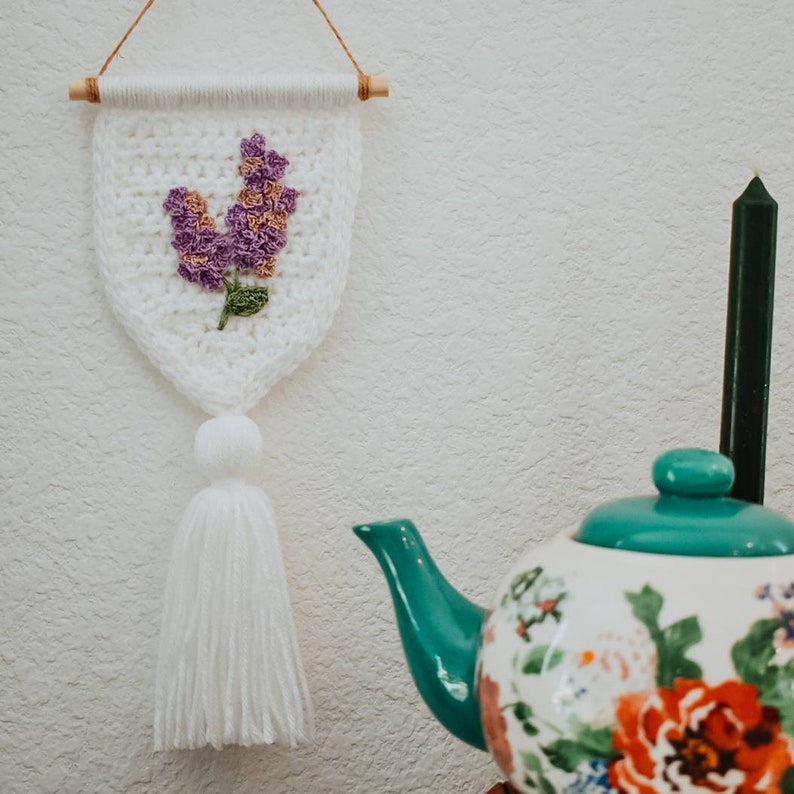 Op de afbeelding: Witte gehaakte wanddecoratie met een paars bloemenmotief en een witte kwast. De decoratie hangt aan een witte muur met een turquoise theepot met bloemenmotieven op de achtergrond.