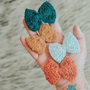 May include: Four crocheted hair bows in different colors and patterns. The bows are teal, light blue, orange, and brown.