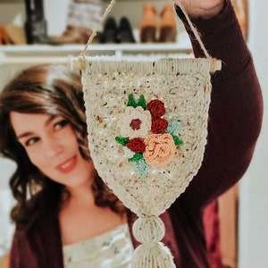 Op de afbeelding: Een witte gehaakte wandhanger met een bloemenontwerp met roze, rode en groene bloemen. De hanger heeft een witte kwast.
