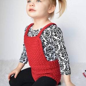 May include: A young child wearing a red crocheted overall with black leggings and a black and white patterned shirt. The child is sitting on a white fluffy surface.