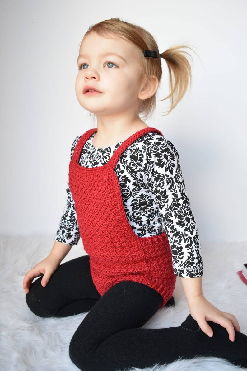 May include: A young child wearing a red crocheted overall with black leggings and a black and white patterned shirt. The child is sitting on a white fluffy surface.