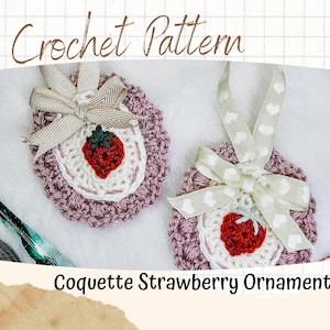 Op de afbeelding: Twee gehaakte aardbei-versieringen met wit en roze garen. Een versiering heeft een bruin en wit lint met een wit hartontwerp. De andere versiering heeft een bruin en wit lint met een witte strik. Beide versieringen hebben een rode gehaakte aardbei in het midden.