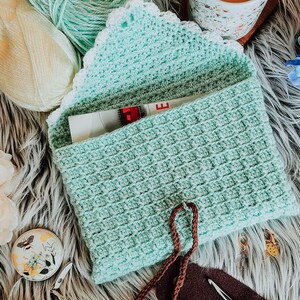 May include: A crocheted teal pouch with a white lace trim and brown drawstrings. The pouch is open and a measuring tape is visible inside. The pouch is sitting on a fluffy white surface with balls of yarn, a cup of tea, and other craft supplies.