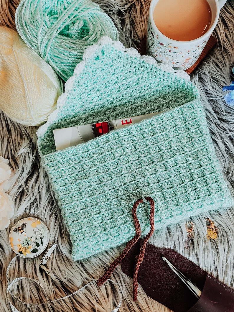 May include: A crocheted teal pouch with a white lace trim and brown drawstrings. The pouch is open and a measuring tape is visible inside. The pouch is sitting on a fluffy white surface with balls of yarn, a cup of tea, and other craft supplies.