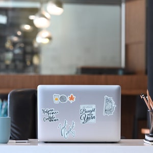 May include: A laptop covered in stickers, including a coffee cup, yarn ball, and maple leaf. Text on the laptop reads "Autumn leaves crochet me", "Just crochet", and "Life update: Bought more yarn". A blue mug and pencils are also on the desk.