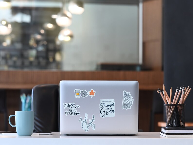 May include: A laptop covered in stickers, including a coffee cup, yarn ball, and maple leaf. Text on the laptop reads "Autumn leaves crochet me", "Just crochet", and "Life update: Bought more yarn". A blue mug and pencils are also on the desk.