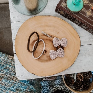 May include: Two small pink crocheted bows with a silver hair clip and two brown and white hair elastics on a wooden surface.
