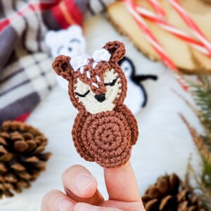 May include: A small, brown and cream-colored crochet deer applique featuring a rounded body, pointed ears, and a face with closed eyes and a black nose. The deer has white stitched details on its forehead and ears, crafted with yarn.