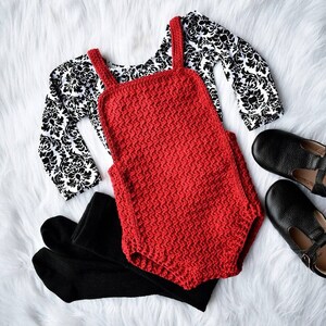 May include: Red crocheted baby romper with black leggings and a black and white patterned long-sleeved shirt. The romper has a simple design with straps and a scoop neck.