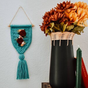 Op de afbeelding: Een turquoise gehaakte wandhanger met een kwast en een bloemenmotief. De hanger wordt getoond op een witte muur met een vaas met herfstbloemen en boeken op de achtergrond.