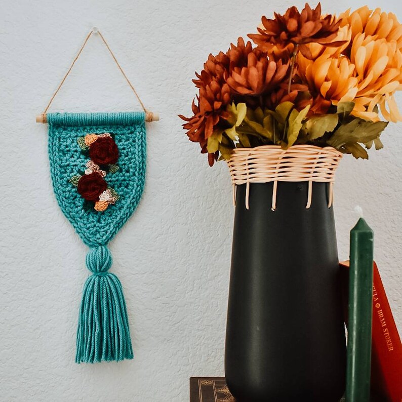 Op de afbeelding: Een turquoise gehaakte wandhanger met een kwast en een bloemenmotief. De hanger wordt getoond op een witte muur met een vaas met herfstbloemen en boeken op de achtergrond.