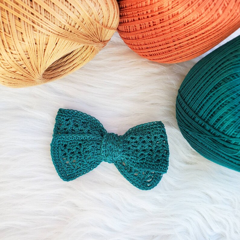 May include: A teal green crocheted bow with a delicate, lacy design. The bow is made with yarn and has a soft, textured appearance.