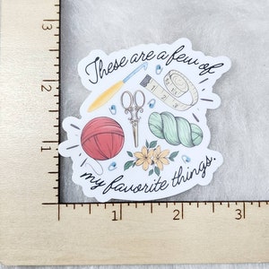 May include: A sticker featuring a collection of crafting tools and supplies. The design includes a red yarn ball, scissors, a crochet hook, a measuring tape, and yarn. The text "These are a few of my favorite things" is also included.