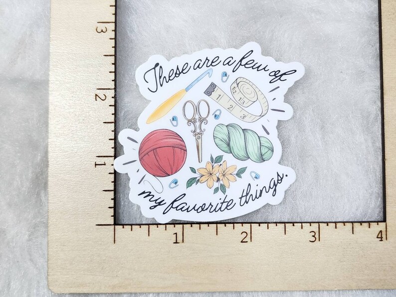 May include: A sticker featuring a collection of crafting tools and supplies. The design includes a red yarn ball, scissors, a crochet hook, a measuring tape, and yarn. The text "These are a few of my favorite things" is also included.
