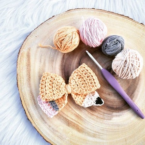 May include: A crocheted bow with a pink, gray, and orange color scheme. The bow is on a wooden surface with balls of yarn in various colors.