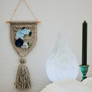 Op de afbeelding: Een beige gehaakte wandhanger met een blauw en groen bloemenontwerp en een kwast. De hanger hangt aan een witte muur naast een witte diffuser en een groene kaars.