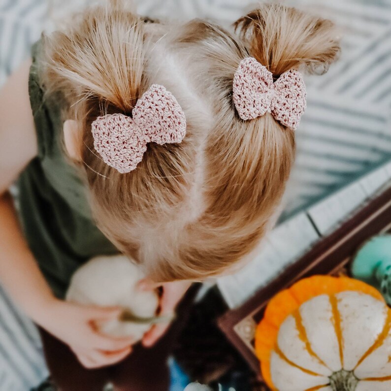 May include: A young girl with blonde hair wearing two pink crocheted bows in her hair.