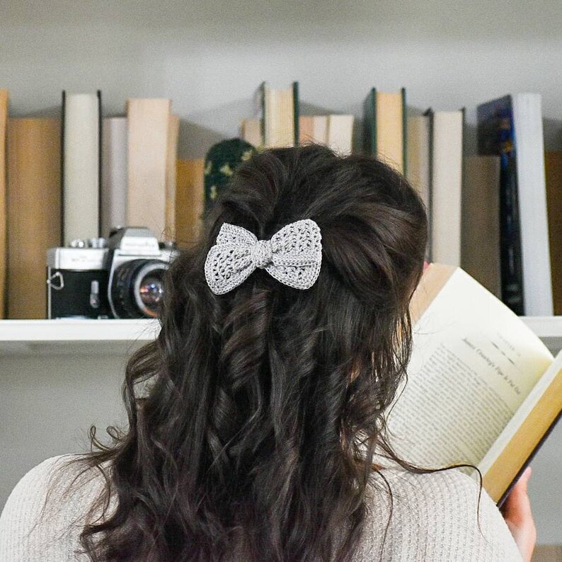 May include: A woman with long, dark brown hair is reading a book. She is wearing a gray sweater and has a gray crocheted bow in her hair.