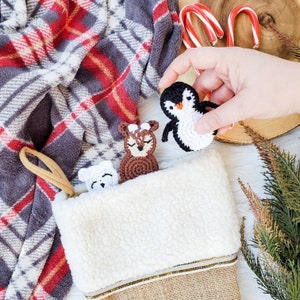 May include: A Christmas stocking with a white faux-shearling cuff and burlap base, containing small crocheted animal figures including a white bear and a brown deer, with a crocheted penguin figure held above the stocking opening.