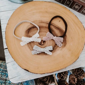 May include: Three hair bows with different colored lace on a wooden surface. The bows are attached to elastic headbands. The headbands are beige, black, and white.