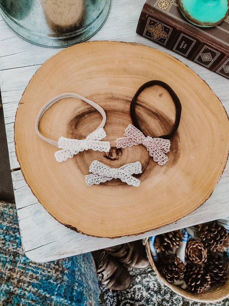 May include: Three hair bows with different colored lace on a wooden surface. The bows are attached to elastic headbands. The headbands are beige, black, and white.