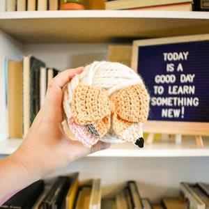 May include: A ball of white yarn with a crocheted fox head attached. The fox head is made of beige, orange, and pink yarn. The yarn ball is being held by a hand.