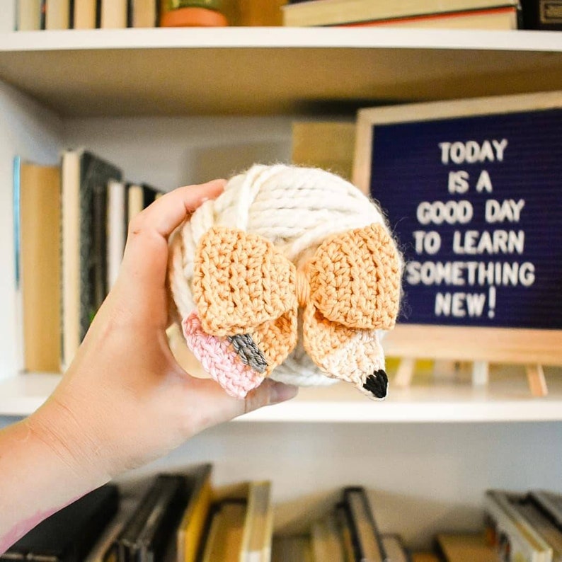 May include: A ball of white yarn with a crocheted fox head attached. The fox head is made of beige, orange, and pink yarn. The yarn ball is being held by a hand.