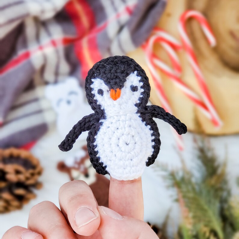 May include: A small, handmade crochet finger puppet shaped like a penguin. The penguin features a black head and back, a white belly and face, two small black eyes, and a bright orange beak. The yarn texture is visible throughout the piece.
