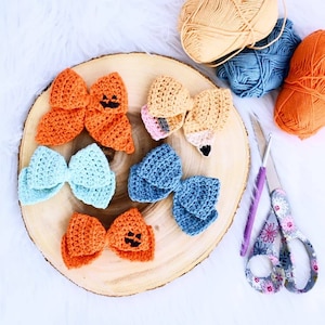 May include: Five crocheted bows on a wooden surface. The bows are in various colors, including orange, blue, and light blue. Two of the orange bows have a black jack-o'-lantern face.