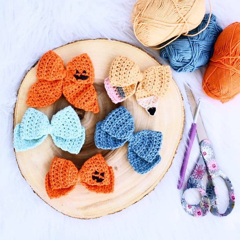 May include: Five crocheted bows on a wooden surface. The bows are in various colors, including orange, blue, and light blue. Two of the orange bows have a black jack-o'-lantern face.