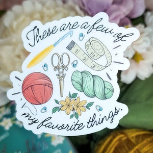 May include: A sticker featuring a collection of crafting tools: a crochet hook, scissors, measuring tape, yarn, and a ball of yarn. The text "These are a few of my favorite things" is written in cursive. The sticker has a white border and is decorated with small flowers.