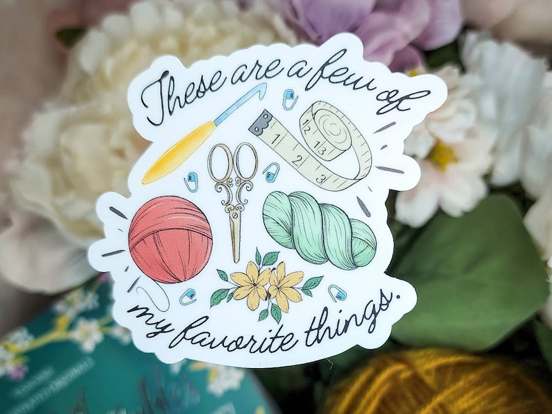May include: A sticker featuring a collection of crafting tools: a crochet hook, scissors, measuring tape, yarn, and a ball of yarn. The text "These are a few of my favorite things" is written in cursive. The sticker has a white border and is decorated with small flowers.
