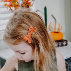 May include: A young girl with long blonde hair wearing a green tank top has a crocheted orange bow in her hair.