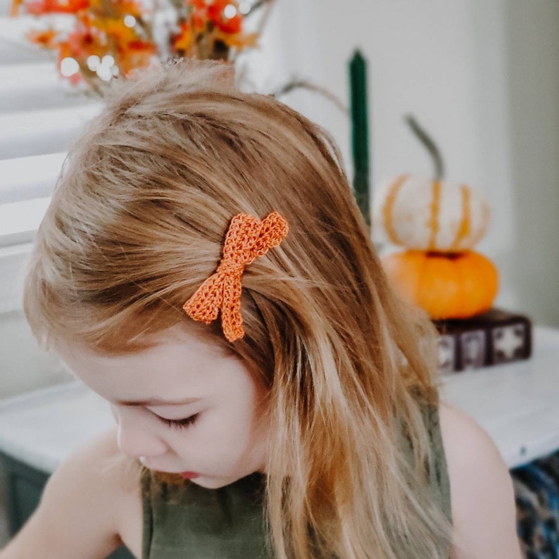 May include: A young girl with long blonde hair wearing a green tank top has a crocheted orange bow in her hair.
