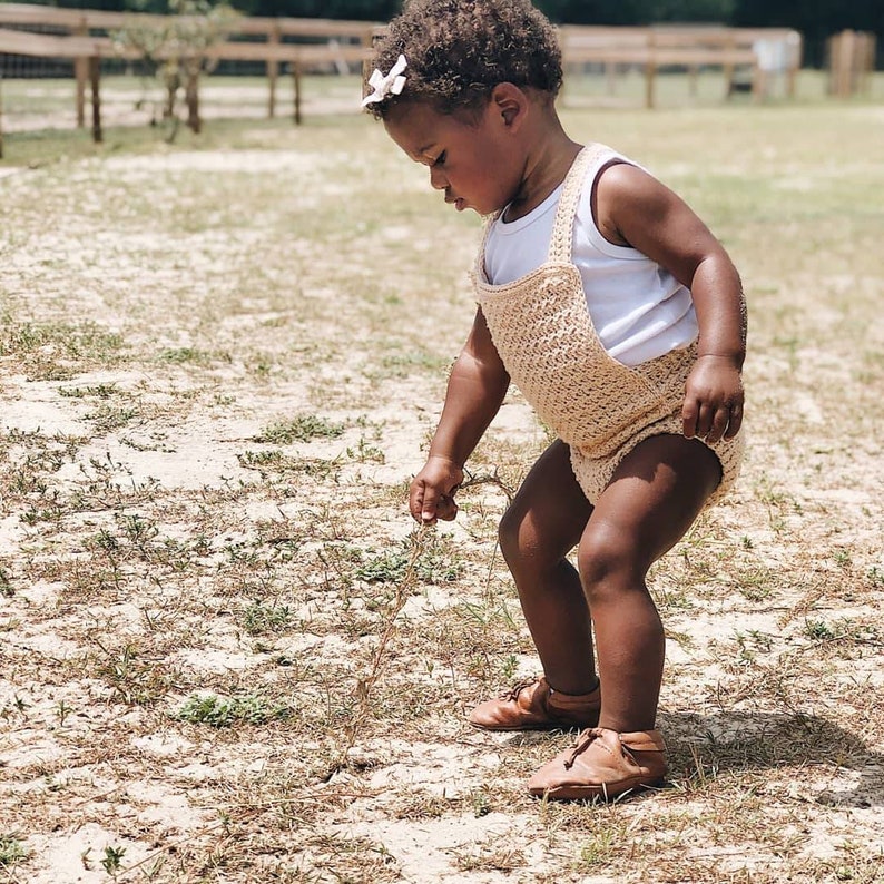 May include: A young child wearing a light brown crocheted romper and brown leather shoes is playing in a grassy field. The child is looking down at the ground and holding a small twig in their hand.