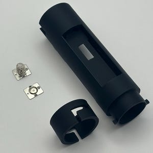 Puede incluir: Una carcasa cilíndrica de plástico negro con un recorte rectangular, un anillo de plástico negro separado con un borde dentado y dos pequeñas placas metálicas cuadradas, cada una con un punto de contacto circular y un resorte en espiral.
