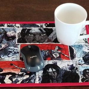 May include: A white ceramic mug sits on a comic book-themed mousepad with a red border. A black Logitech mouse sits on the mousepad. The mousepad features a comic book panel design with red, black, and grey tones.