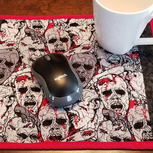 May include: A black and grey computer mouse sits on a red-bordered mouse mat with a pattern of zombie faces. The mouse mat is on a wooden surface.