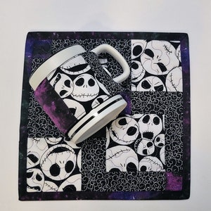 Puede incluir: Una taza blanca y negra con un patr&oacute;n del rostro de Jack Skellington de la pel&iacute;cula Pesadilla antes de Navidad. La taza est&aacute; sobre un posavasos de tela blanco y negro con un patr&oacute;n similar. El posavasos tiene un borde morado y negro.