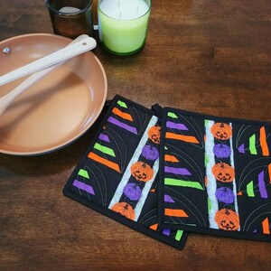 May include: Two black Halloween-themed pot holders with orange, green and purple stripes and jack-o'-lantern designs.