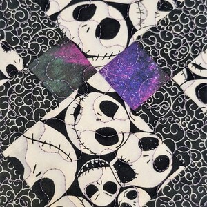 Puede incluir: Un posavasos de tela blanco y negro con un patr&oacute;n repetido de la cara de Jack Skellington. El posavasos tiene un borde morado y negro con un patr&oacute;n de galaxia.