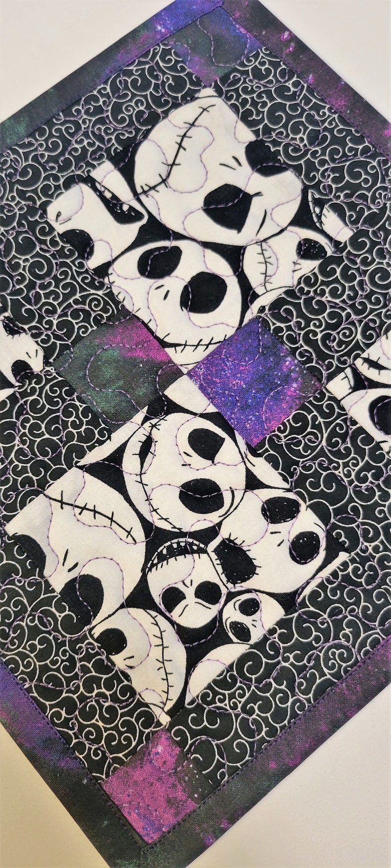 Puede incluir: Un posavasos de tela blanco y negro con un patr&oacute;n repetido de la cara de Jack Skellington. El posavasos tiene un borde morado y negro con un patr&oacute;n de galaxia.