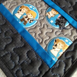 K&ouml;nnte beinhalten: Ein schwarzer und grauer gesteppter Untersetzer mit einem blauen Bandrand. Der Untersetzer zeigt ein sich wiederholendes Muster von Comicfiguren aus Star Wars, darunter Prinzessin Leia, Han Solo und Darth Vader.