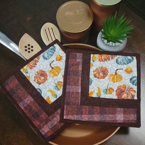 May include: Two brown quilted pot holders with a pumpkin and gourd pattern. The pot holders have a brown and red plaid border.