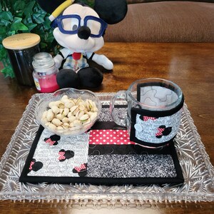 May include: A glass mug with a fabric Minnie Mouse themed cup cozy sits on a glass tray with a bowl of pistachios and a Minnie Mouse themed fabric coaster.