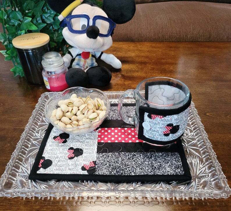 May include: A glass mug with a fabric Minnie Mouse themed cup cozy sits on a glass tray with a bowl of pistachios and a Minnie Mouse themed fabric coaster.