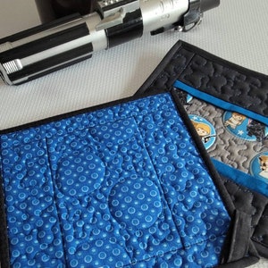 K&ouml;nnte beinhalten: Zwei blaue und schwarze gequiltete Topflappen mit einem wei&szlig;en Etikett, auf dem "Lightside Creations" steht. Ein Topflappen hat einen blauen Hintergrund mit wei&szlig;en Punkten und der andere einen grauen Hintergrund mit einem Star Wars-Charakterdruck.
