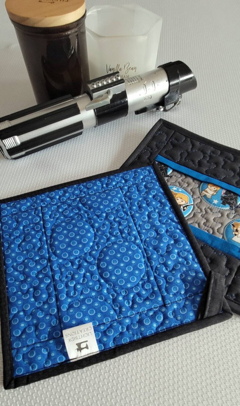 K&ouml;nnte beinhalten: Zwei blaue und schwarze gequiltete Topflappen mit einem wei&szlig;en Etikett, auf dem "Lightside Creations" steht. Ein Topflappen hat einen blauen Hintergrund mit wei&szlig;en Punkten und der andere einen grauen Hintergrund mit einem Star Wars-Charakterdruck.